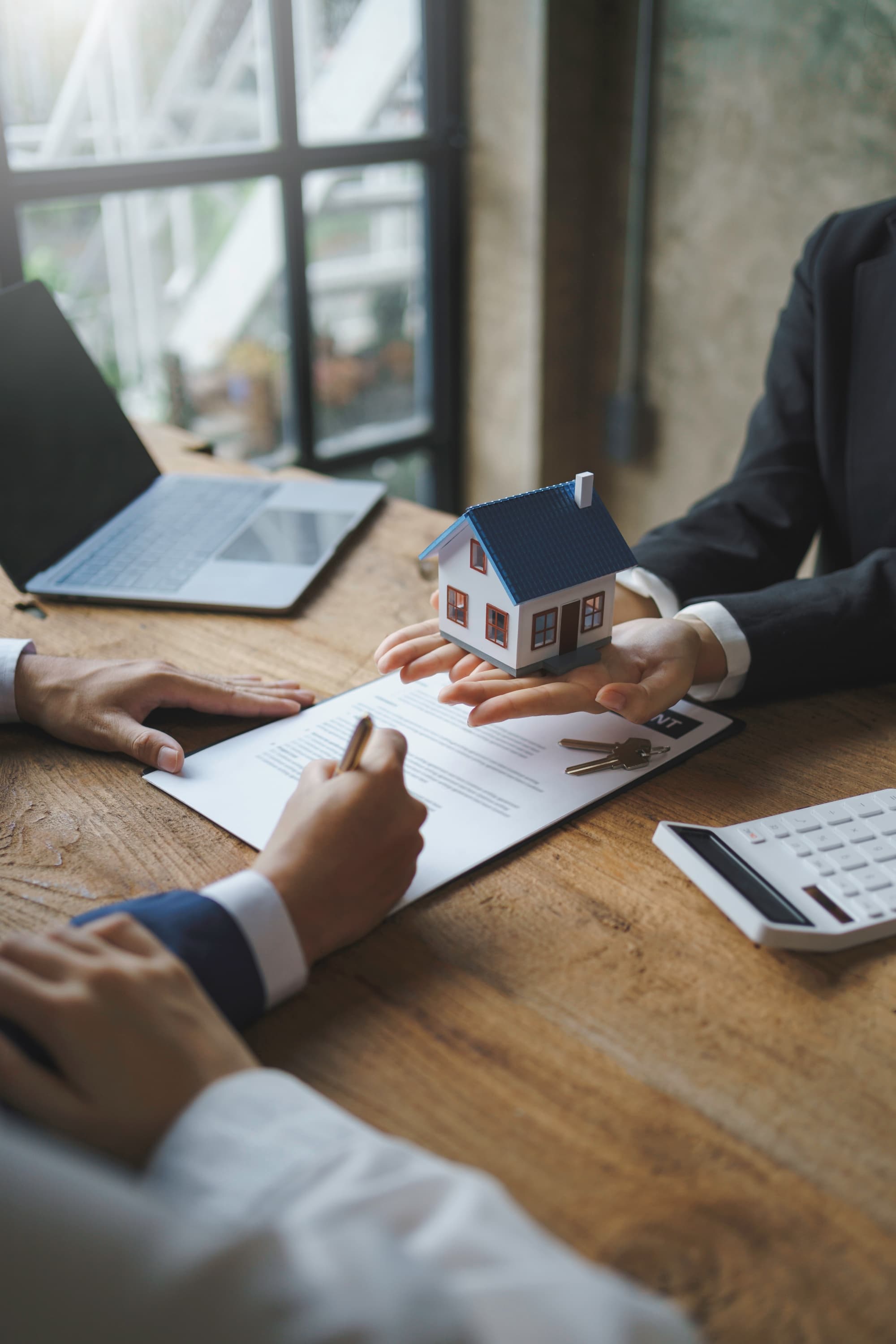 Two professionals reviewing a real estate contract with a miniature house model and property keys on a desk