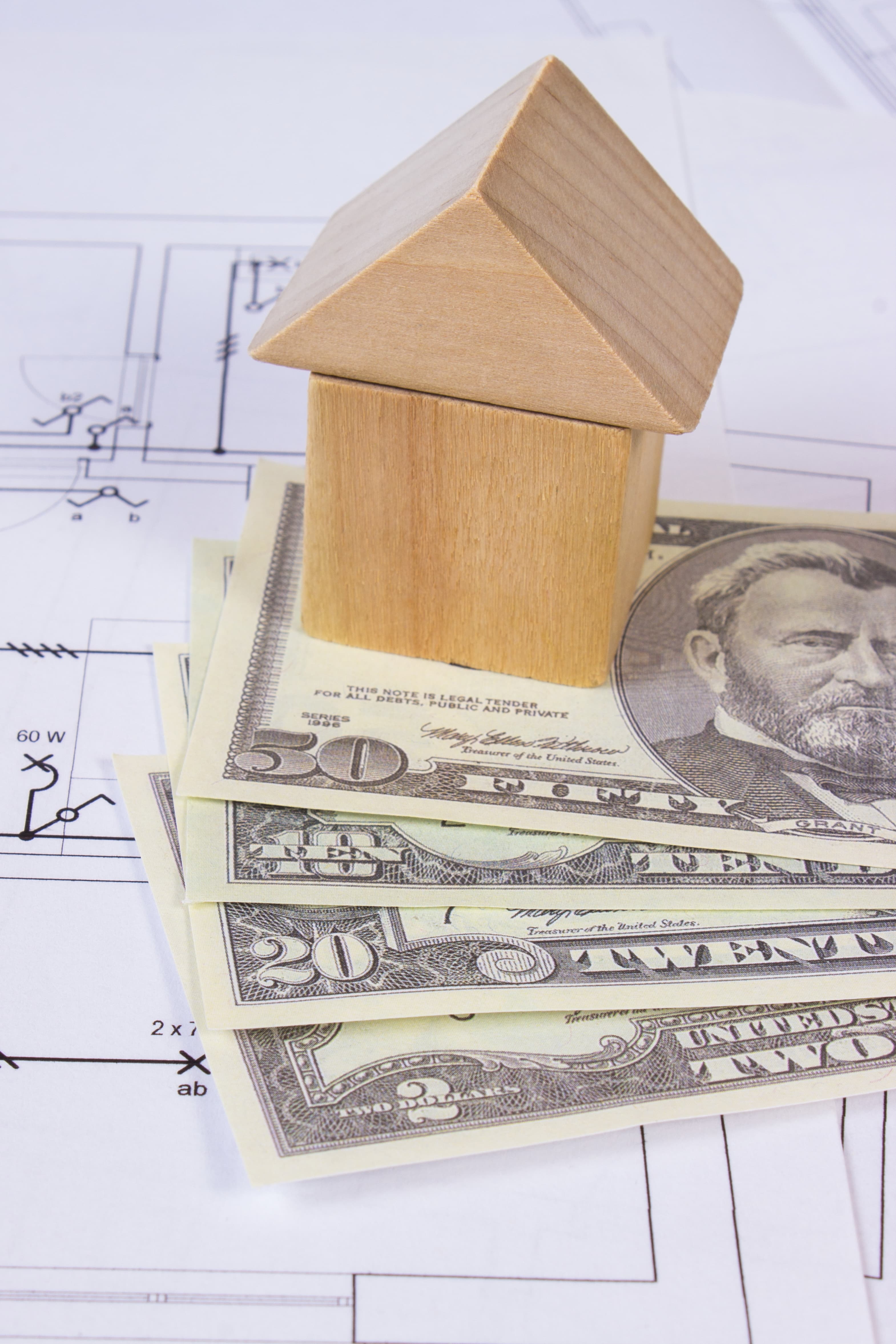 Wooden house figurine resting on US dollar bills laid over architectural blueprints